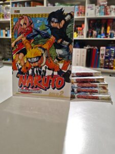 Um volume da edição Naruto Gold em pé na frente, com outros volumes empilhados atrás mostrando as laterais com os títulos e números das edições, destacando o acabamento diferenciado e a arte de Masashi Kishimoto na capa.