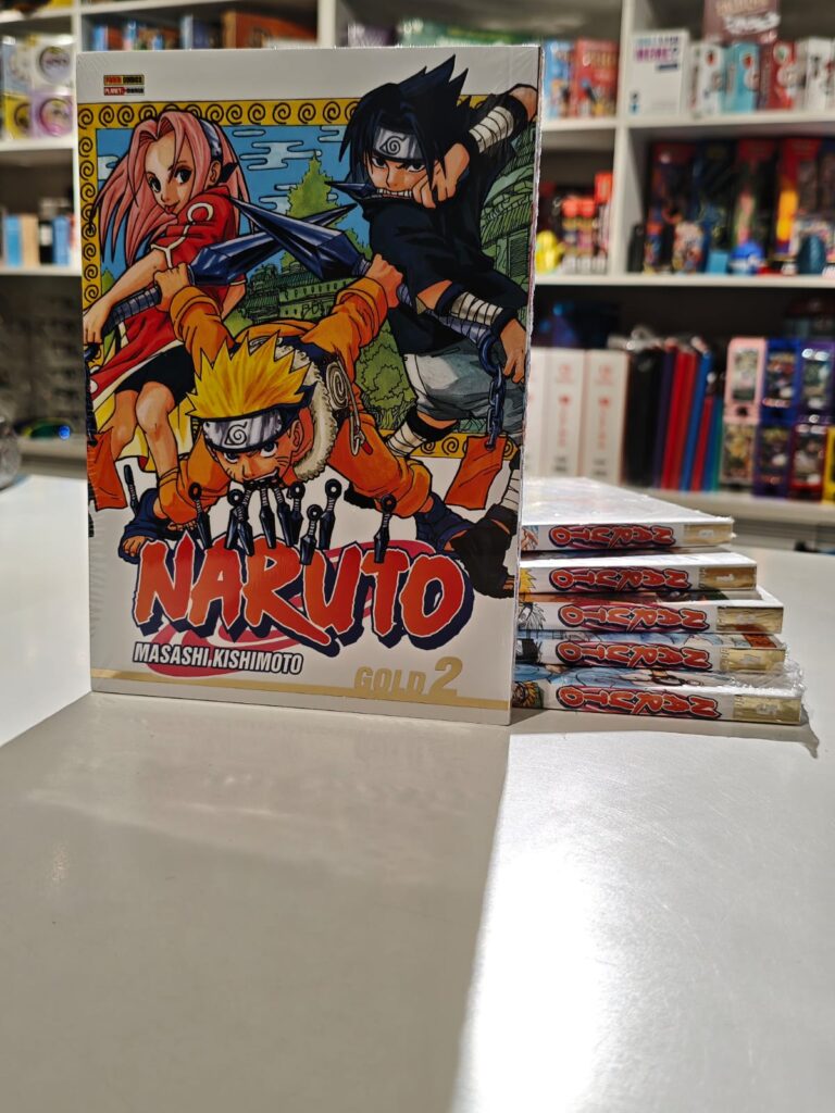 Um volume da edição Naruto Gold em pé na frente, com outros volumes empilhados atrás mostrando as laterais com os títulos e números das edições, destacando o acabamento diferenciado e a arte de Masashi Kishimoto na capa.