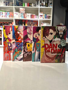 Cinco primeiros volumes do mangá Dandadan enfileirados em pé, mostrando as capas sequenciais com os protagonistas Momo e Okarun em poses dinâmicas e elementos sobrenaturais ao fundo, arte característica de Yukinobu Tatsu.