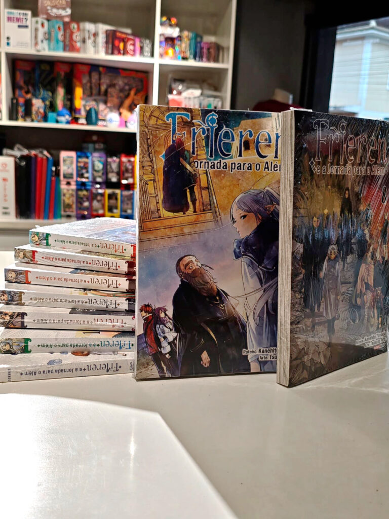 Dois primeiros volumes do mangá Frieren e a Jornada para o Além em pé na frente, com outros volumes empilhados atrás mostrando as laterais com os títulos e números das edições, destacando a arte de Tsukasa Abe com a personagem Frieren.